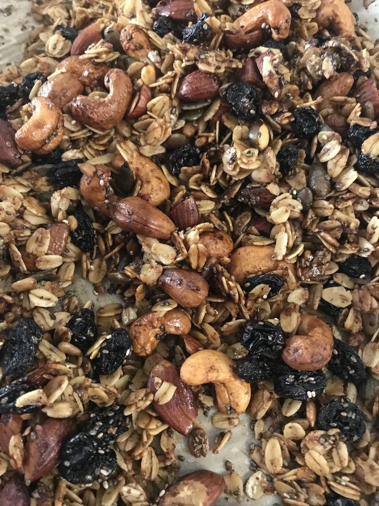 golden brown glistening roasted homemade granola spread out on baking sheet over white parchment paper, cashew nuts, chopped almonds, raisins, rolled oats